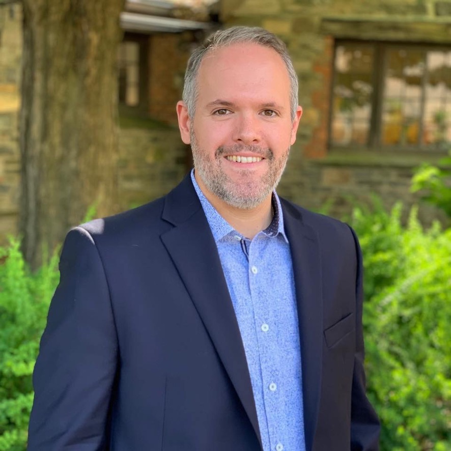 Zachary Cohn smiling outdoors in a navy suit